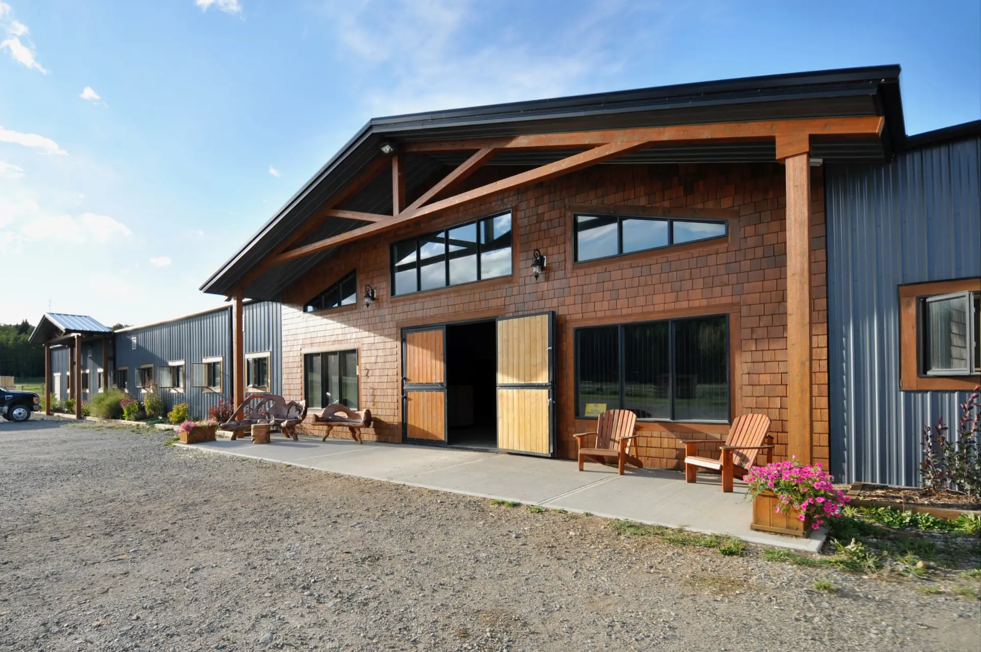 Steel-clad covered riding area with a cedar-shingled lounge entry, timber-framed canopy, sliding doors and Adirondack chairs on the patio