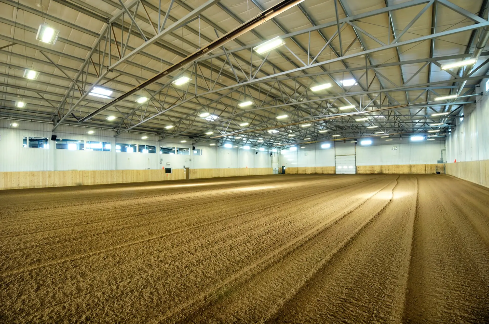 Interior of a clear-span steel riding arena with a groomed sand floor, open steel roof trusses, LED lights and a timber kick wall