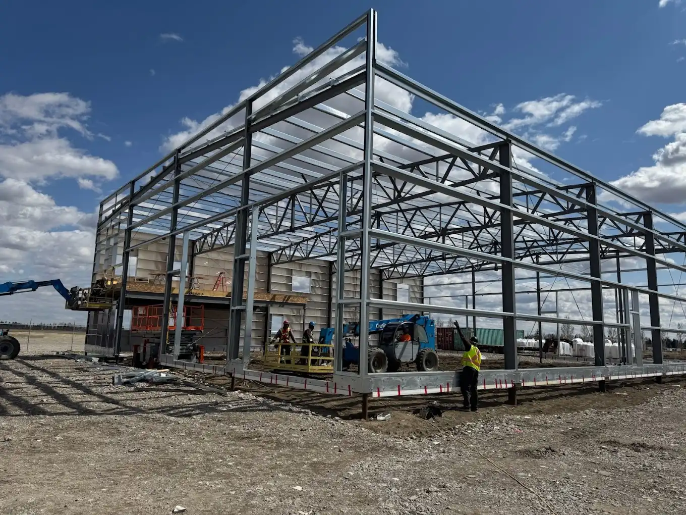 Two-storey steel building frame going up on an Alberta site, with crews working from a telehandler basket and roof joists already set
