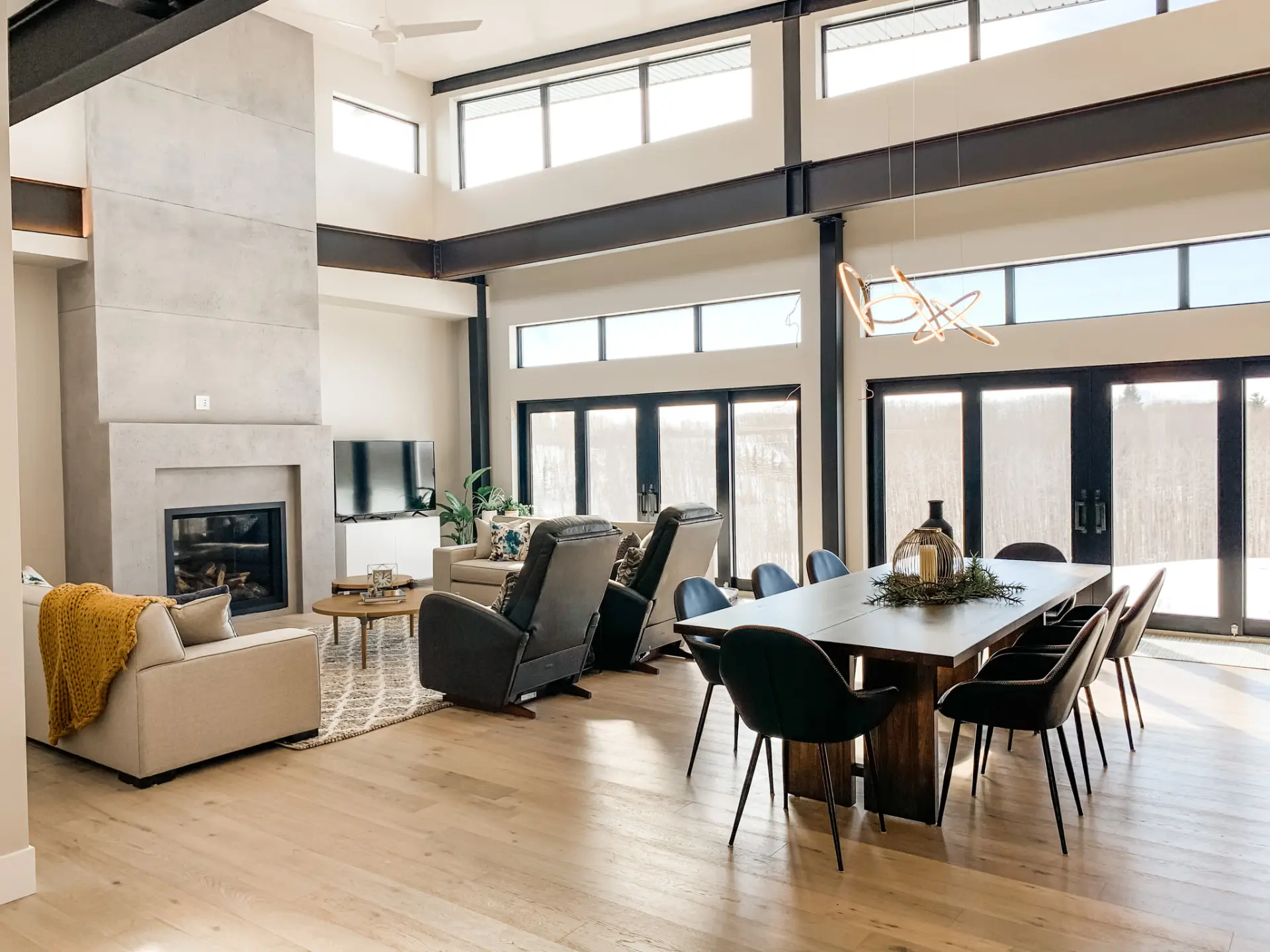 Open-plan great room with exposed dark steel beams, clerestory windows, full-height glass doors, a concrete fireplace wall and a dining table