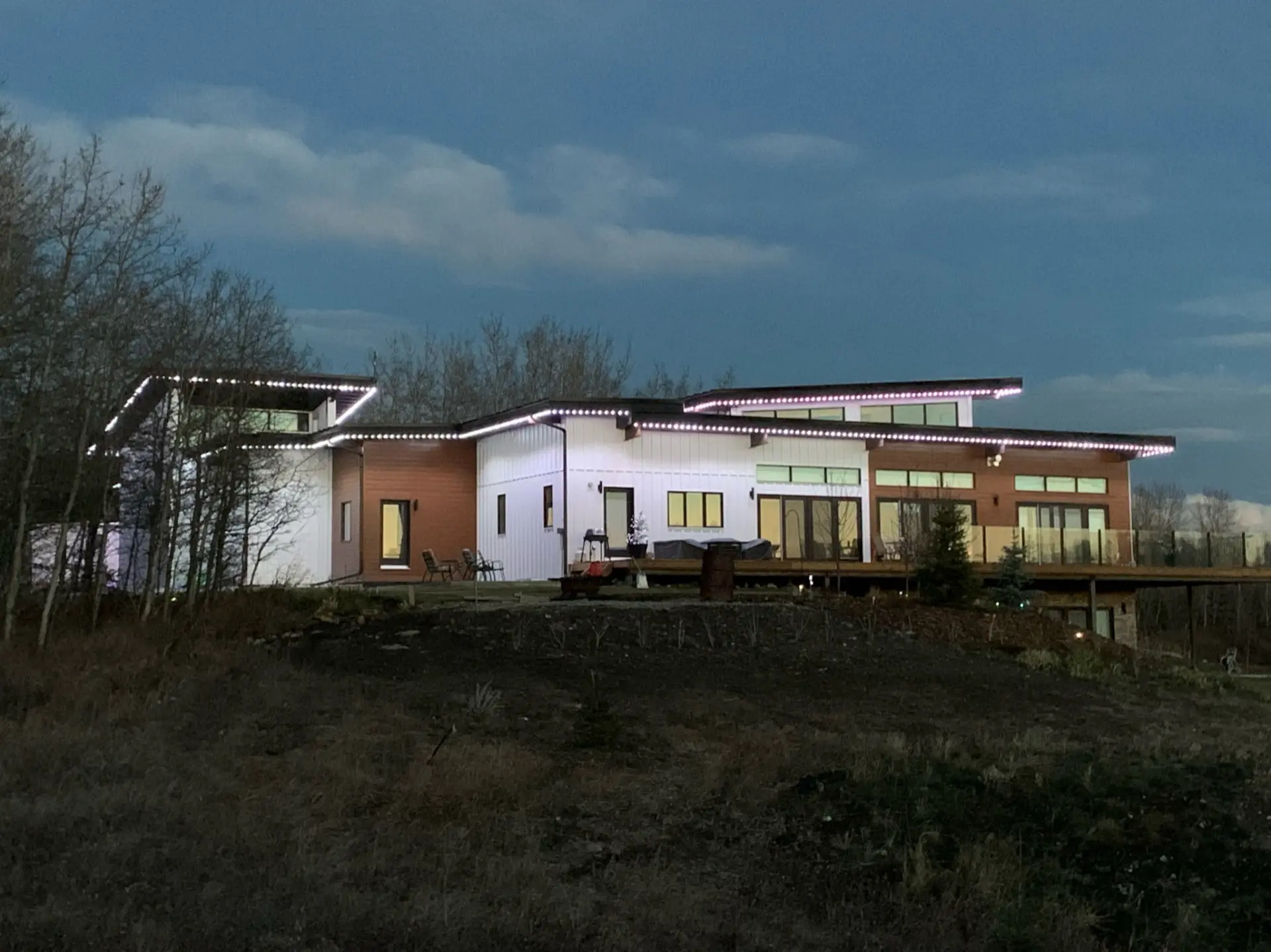 Modern steel-frame home at dusk with white board-and-batten and wood-tone cladding, a long raised deck with glass railing, and roofline lighting