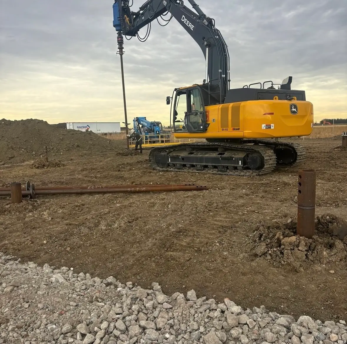 Excavator-mounted drive installing a steel pile on the commercial tire shop site in Rocky View County, with finished piles in the foreground
